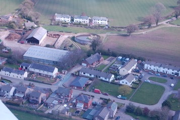 An aerial view of a rural area featuring residential buildings with pitched roofs arranged in orderly rows and clusters. A large barn and several agricultural structures are visible, along with open fields and grassy areas. The scene is surrounded by trees and shrubs, suggesting a countryside setting with a mix of modern and traditional architecture.