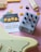 A collection of music-related items arranged on a light pink surface. In the center is a silver guitar effects pedal labeled 'Draume' with various knobs for control. To the left, a set of yellow guitar picks is placed on a card with details about the pick type, labeled 'Rombo Jade' with a thickness of 2.3 mm. A portion of a cream-colored electric guitar is visible in the lower part of the image. The overall composition includes additional scattered guitar picks and pickguards.