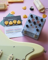 A collection of music-related items arranged on a light pink surface. In the center is a silver guitar effects pedal labeled 'Draume' with various knobs for control. To the left, a set of yellow guitar picks is placed on a card with details about the pick type, labeled 'Rombo Jade' with a thickness of 2.3 mm. A portion of a cream-colored electric guitar is visible in the lower part of the image. The overall composition includes additional scattered guitar picks and pickguards.