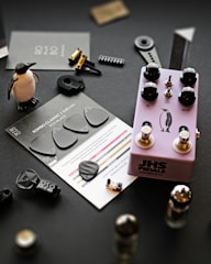 A collection of music-related items and small objects is arranged on a dark surface. A purple guitar effects pedal labeled 'JHS Pedals' with black knobs sits centrally, featuring a penguin graphic. Nearby, black guitar picks are displayed on a card labeled 'ROMBO CLASSIC, 0.45mm, ECO BLACK'. A small penguin figurine stands upright. Various mechanical components, including metallic parts and tiny plastic pieces, are scattered around.