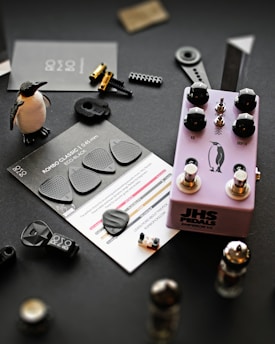A collection of music-related items and small objects is arranged on a dark surface. A purple guitar effects pedal labeled 'JHS Pedals' with black knobs sits centrally, featuring a penguin graphic. Nearby, black guitar picks are displayed on a card labeled 'ROMBO CLASSIC, 0.45mm, ECO BLACK'. A small penguin figurine stands upright. Various mechanical components, including metallic parts and tiny plastic pieces, are scattered around.