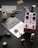A collection of music-related items and small objects is arranged on a dark surface. A purple guitar effects pedal labeled 'JHS Pedals' with black knobs sits centrally, featuring a penguin graphic. Nearby, black guitar picks are displayed on a card labeled 'ROMBO CLASSIC, 0.45mm, ECO BLACK'. A small penguin figurine stands upright. Various mechanical components, including metallic parts and tiny plastic pieces, are scattered around.