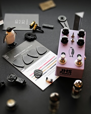 A collection of music-related items and small objects is arranged on a dark surface. A purple guitar effects pedal labeled 'JHS Pedals' with black knobs sits centrally, featuring a penguin graphic. Nearby, black guitar picks are displayed on a card labeled 'ROMBO CLASSIC, 0.45mm, ECO BLACK'. A small penguin figurine stands upright. Various mechanical components, including metallic parts and tiny plastic pieces, are scattered around.