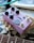 A purple guitar effects pedal with several black knobs and switches, featuring an illustration of a penguin and a fish. The pedal rests on a mannequin hand above a black and white patterned surface. Additional items, like a guitar neck and an open book, are visible in the background.