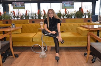 A woman seated on a mustard yellow couch is holding a hookah in a modern lounge setting. The room has large windows with potted plants and there are televisions mounted on the wall displaying sports. The wooden floor and light-colored tables create a cozy atmosphere.