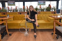 A woman seated on a mustard yellow couch is holding a hookah in a modern lounge setting. The room has large windows with potted plants and there are televisions mounted on the wall displaying sports. The wooden floor and light-colored tables create a cozy atmosphere.