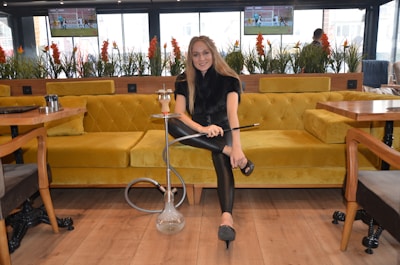 A woman seated on a mustard yellow couch is holding a hookah in a modern lounge setting. The room has large windows with potted plants and there are televisions mounted on the wall displaying sports. The wooden floor and light-colored tables create a cozy atmosphere.