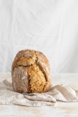 Close-up of a rustic bread loaf with a crisp crust on a white plate against a simple beige background.