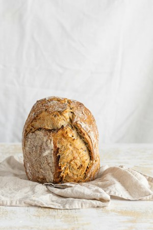 A rustic, round loaf of bread with a golden, crispy crust is placed on a lightly colored cloth. The background is simple and neutral.