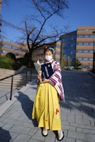 A person in traditional attire is holding a diploma and a bouquet of flowers. The outfit consists of a yellow skirt with a patterned shawl over it. The background features a modern building and a bare tree under a clear blue sky.