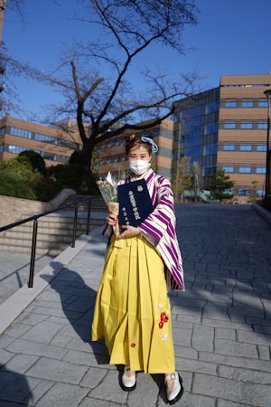 A person in traditional attire is holding a diploma and a bouquet of flowers. The outfit consists of a yellow skirt with a patterned shawl over it. The background features a modern building and a bare tree under a clear blue sky.