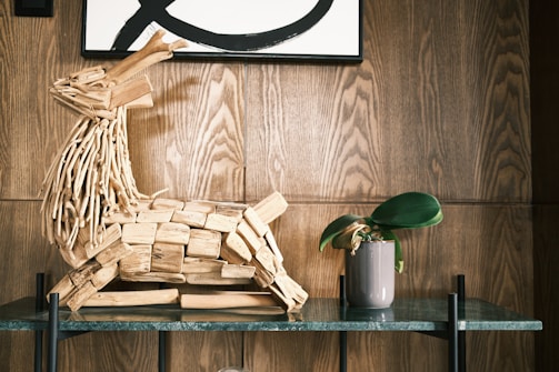 A wooden sculpture made from driftwood pieces resembling an animal with a long neck sits on a glass shelf. Next to it is a small potted plant with wide green leaves. The background features a wooden panel wall and part of a framed artwork with bold black lines.