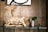 A wooden sculpture made from driftwood pieces resembling an animal with a long neck sits on a glass shelf. Next to it is a small potted plant with wide green leaves. The background features a wooden panel wall and part of a framed artwork with bold black lines.