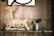 A wooden sculpture made from driftwood pieces resembling an animal with a long neck sits on a glass shelf. Next to it is a small potted plant with wide green leaves. The background features a wooden panel wall and part of a framed artwork with bold black lines.