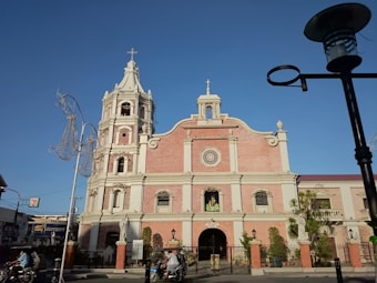 An ornate and historic church with red brick and stone architecture stands prominently under a clear blue sky. A tower with a cross sits on the left side, and statues decorate the façade. Nearby, a few people are seen on a street with motorcycles and other urban elements like street lamps, suggesting a vibrant surrounding.