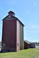A rustic, red barn-like structure stands prominently on a grassy area, adjacent to several tall, cylindrical silos. The building has a weathered appearance with small windows near the top. The sky is clear and blue, adding to the open, rural setting.