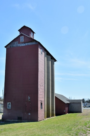A rustic, red barn-like structure stands prominently on a grassy area, adjacent to several tall, cylindrical silos. The building has a weathered appearance with small windows near the top. The sky is clear and blue, adding to the open, rural setting.