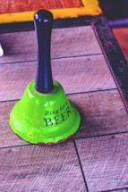 A vintage green bell with a black handle rests on a wooden surface. The bell has the phrase 'Ring for a BEER' written on it in bold, black letters. The texture of the wood is visible, and the bell shows signs of wear with spots of rustic discoloration along its rim.