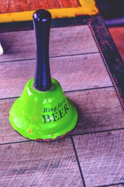 A vintage green bell with a black handle rests on a wooden surface. The bell has the phrase 'Ring for a BEER' written on it in bold, black letters. The texture of the wood is visible, and the bell shows signs of wear with spots of rustic discoloration along its rim.