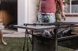 Outdoor scene with a barbecue grill loaded with charcoal and partially charred wood. A person is standing nearby, wearing a pink long-sleeve shirt and striped shorts. Another figure is partially visible on the left side.