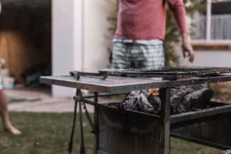 Outdoor scene with a barbecue grill loaded with charcoal and partially charred wood. A person is standing nearby, wearing a pink long-sleeve shirt and striped shorts. Another figure is partially visible on the left side.