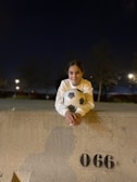 A person dressed in a white sweater is leaning on a concrete barrier, holding a soccer ball. It is nighttime, with streetlights illuminating the background, creating a calm and quiet ambience. Shadows are cast on the concrete wall, and the number 066 is stenciled on it.
