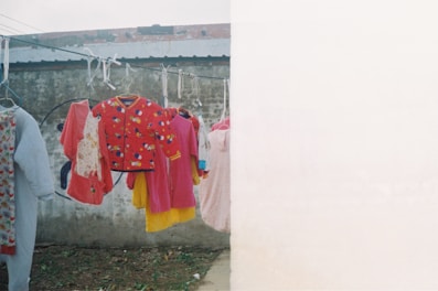 Clothes are hanging on a laundry line against a weathered concrete wall. Various colorful garments including a red jacket with cartoon patterns, a pink dress, and other children's clothing are prominently displayed.