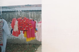 Clothes are hanging on a laundry line against a weathered concrete wall. Various colorful garments including a red jacket with cartoon patterns, a pink dress, and other children's clothing are prominently displayed.