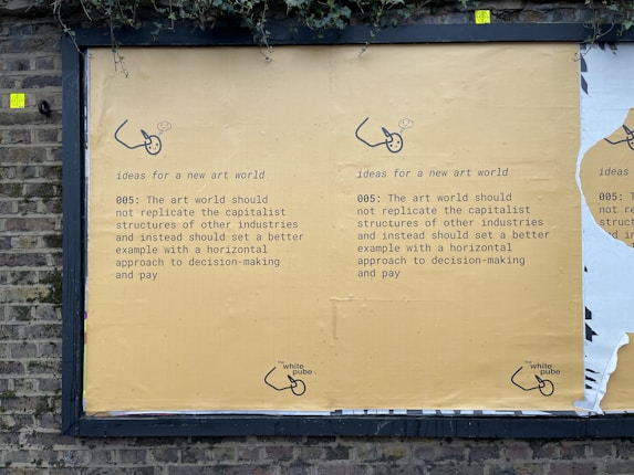 A street mural on a brick wall displays a message advocating for change in the art world. It suggests not replicating capitalist structures and instead promoting horizontal decision-making and equitable pay. The mural is mainly yellow with black text and features a small logo or illustration at the bottom.