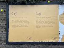 A street mural on a brick wall displays a message advocating for change in the art world. It suggests not replicating capitalist structures and instead promoting horizontal decision-making and equitable pay. The mural is mainly yellow with black text and features a small logo or illustration at the bottom.