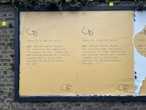 A street mural on a brick wall displays a message advocating for change in the art world. It suggests not replicating capitalist structures and instead promoting horizontal decision-making and equitable pay. The mural is mainly yellow with black text and features a small logo or illustration at the bottom.