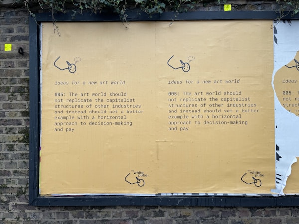 A street mural on a brick wall displays a message advocating for change in the art world. It suggests not replicating capitalist structures and instead promoting horizontal decision-making and equitable pay. The mural is mainly yellow with black text and features a small logo or illustration at the bottom.