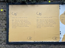 A street mural on a brick wall displays a message advocating for change in the art world. It suggests not replicating capitalist structures and instead promoting horizontal decision-making and equitable pay. The mural is mainly yellow with black text and features a small logo or illustration at the bottom.