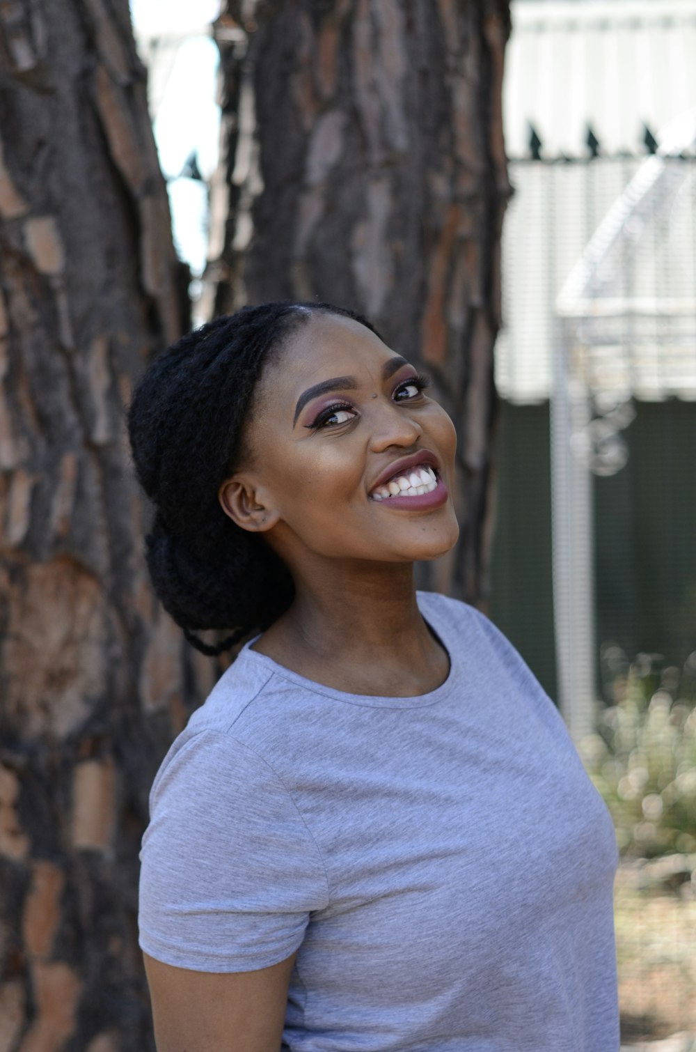 smiling woman in gray crew neck shirt standing beside brown tree during daytime