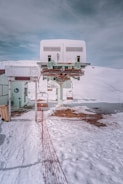 A ski lift station located on a snowy mountain, with a cable and pulley system overhead. There is a red safety net fence lining the path leading up to the lift station. The surrounding area is covered with snow, and the sky above is overcast with gray clouds. A partially visible sign with instructions is present near the building.