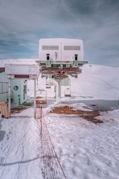 A ski lift station located on a snowy mountain, with a cable and pulley system overhead. There is a red safety net fence lining the path leading up to the lift station. The surrounding area is covered with snow, and the sky above is overcast with gray clouds. A partially visible sign with instructions is present near the building.