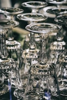 A collection of upside-down wine glasses with intricate stems arranged closely together. The glassware reflects light, creating shimmering highlights and a sense of elegance.