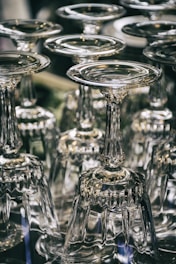 A collection of upside-down wine glasses with intricate stems arranged closely together. The glassware reflects light, creating shimmering highlights and a sense of elegance.