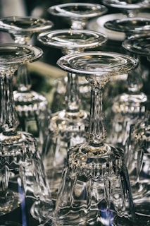 A collection of upside-down wine glasses with intricate stems arranged closely together. The glassware reflects light, creating shimmering highlights and a sense of elegance.