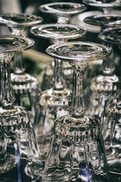 A collection of upside-down wine glasses with intricate stems arranged closely together. The glassware reflects light, creating shimmering highlights and a sense of elegance.
