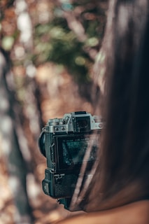 A person is holding a DSLR camera, focusing on capturing a scene. The background is blurred with natural colors, suggesting an outdoor setting.