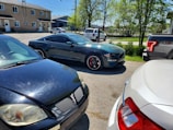 Several cars are parked on a sunny day in a small parking lot surrounded by trees and a few buildings. A black sports car with red brake calipers is prominently featured in the center, flanked by other vehicles including a black sedan and a white truck.