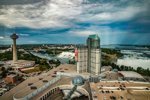 A sprawling urban landscape with a prominent tower and a large building with a green roof. In the background, a river flows over a waterfall, surrounded by lush greenery. The sky is partly cloudy, creating a dramatic atmosphere over the scene.