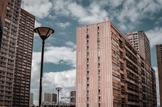 Several high-rise apartment buildings with a modern architectural style are set against a backdrop of a partly cloudy sky. Two street lamps are visible in the foreground, contrasting with the towering structures.