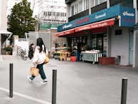 A small urban grocery store named Amasia with blue and red signage, featuring fruits and vegetables displayed outside. Two people are walking on the sidewalk, one carrying a shopping bag. There are bicycles parked nearby and trees in the background.