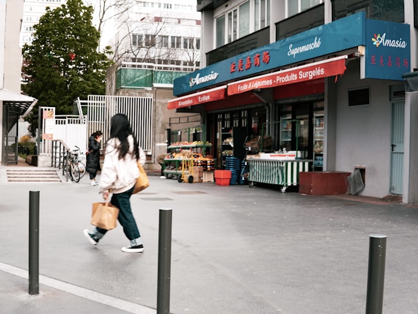 A small urban grocery store named Amasia with blue and red signage, featuring fruits and vegetables displayed outside. Two people are walking on the sidewalk, one carrying a shopping bag. There are bicycles parked nearby and trees in the background.