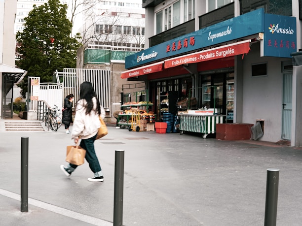 A small urban grocery store named Amasia with blue and red signage, featuring fruits and vegetables displayed outside. Two people are walking on the sidewalk, one carrying a shopping bag. There are bicycles parked nearby and trees in the background.