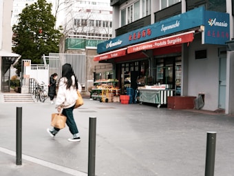 A small urban grocery store named Amasia with blue and red signage, featuring fruits and vegetables displayed outside. Two people are walking on the sidewalk, one carrying a shopping bag. There are bicycles parked nearby and trees in the background.