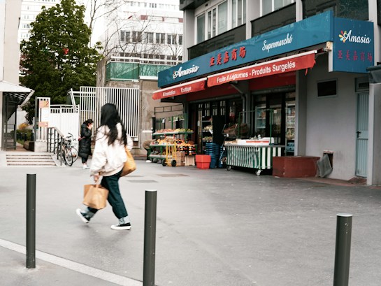 A small urban grocery store named Amasia with blue and red signage, featuring fruits and vegetables displayed outside. Two people are walking on the sidewalk, one carrying a shopping bag. There are bicycles parked nearby and trees in the background.