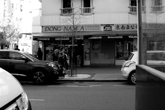 A black and white street scene featuring a storefront with signs reading 'Dong Nam' and 'Produits Exotiques'. Several cars are parked on the street and a few pedestrians can be seen walking past the shop. The architecture of the building and the vehicles suggest an urban setting.